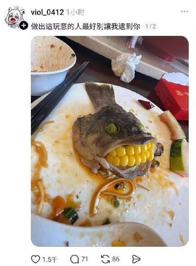 这是一张社交媒体动态截图，发布者账号为viol_0412，配文为“做出這玩意的人最好別讓我逮到你”。截图的实拍内容为用餐后剩余的餐盘，盘中的熟鱼头被人恶搞改造：眼眶处被塞入青豆当作眼睛，鱼嘴中被塞进一段玉米段，看起来仿佛鱼头长了一排整齐的黄色牙齿，造型怪异又充满喜感。餐盘上还残留有酱汁、粉条、葱段，旁边放着筷子、吃剩的西瓜皮，背景可见空碗、用过的纸巾和红色包装盒，整体是日常用餐场景下的趣味整活，反差感十足。