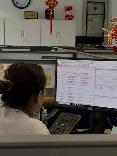 办公室工位场景：一位扎着丸子头的女性职员正专注于电脑屏幕，右侧显示器显示着某公司项目规划方案文档。工位上方的柜子摆放着荣誉证书、红色福字挂饰、生肖鼠玩偶等新春装饰，墙上时钟指向约10:10，整体氛围忙碌又带有节日气息。