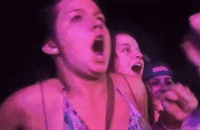 一位女性在演唱会现场表现出极度兴奋的状态，她仰头张嘴呐喊，双臂高高举起，身体随着音乐或现场气氛摆动。周围还有其他观众也在欢呼，整体场景充满了热烈欢快的氛围，紫色的舞台灯光照亮了她激动的表情。
