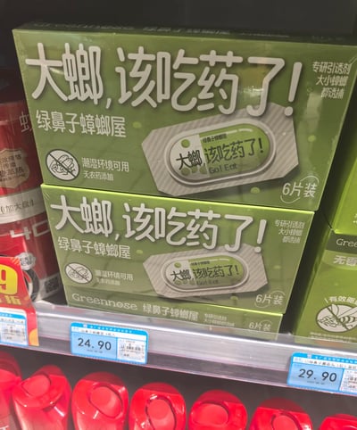 超市货架上摆放着两盒绿色包装的“绿鼻子蟑螂屋”产品，包装正面印有醒目文字“大螂，该吃药了！”，下方配有产品实物图。包装上标注“专研引诱剂 大小蟑螂都诱捕”“潮湿环境可用”“无农药添加”等产品特点，以及“6片装”和品牌名“Greennose 绿鼻子蟑螂屋”。货架标签显示价格分别为24.90元和29.90元，周围可见部分红色包装的其他商品。
