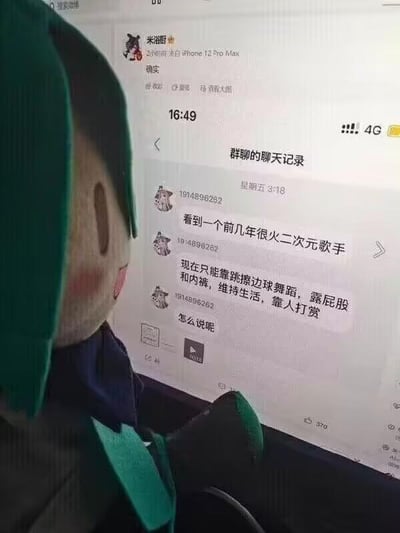 图片中展示了一个绿色头发、蓝色衣服的卡通玩偶正拿着手机，手机屏幕上显示着一段群聊记录。聊天内容提到一位前几年很火的二次元歌手如今只能靠跳擦边球舞蹈、穿内裤跳舞露屁股来维持生活，靠人打赏。玩偶面无表情地注视着这段对话，形成一种微妙的对比。