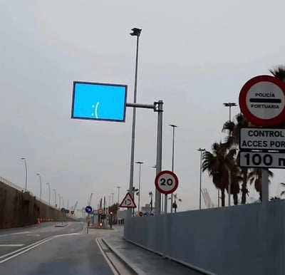 阴天的道路场景，画面中央的电子交通指示牌显示蓝色背景和一个':('悲伤表情符号，周围有多个交通标志，包括红色圆形的'POLICÍA PORTUARIA'（港口警察）标志、'CONTROL ACCES POR 100 m'（100米处进入控制）以及限速20的标识。道路旁有路灯和棕榈树，整体氛围因指示牌的表情显得略带幽默。
