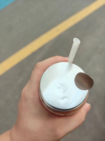 这是一张实拍生活照，画面中一只手握着一罐带铝制顶盖的罐装饮品，顶盖未正常拉开拉环，仅在表面戳孔插入了一根白色塑料吸管；顶盖自然的凹陷痕迹与拉环掀开后留下的椭圆形缺口组合在一起，视觉上酷似一张带有眼睛和嘴巴的人脸，背景为灰色路面，上面有一条斜向的黄色标线。