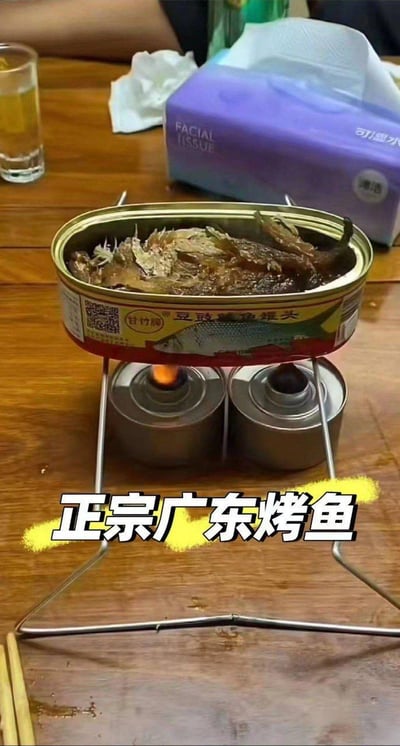 一张餐桌上的搞笑场景图：一个豆豉鲮鱼罐头被架在两个小型燃气灶上加热，罐头里的鱼肉清晰可见。桌子上还放着一杯黄色饮品、一包紫色包装的纸巾和一些纸巾碎屑。图片底部有醒目的黄色高亮文字'正宗广东烤鱼'，用夸张的方式调侃这种简易加热罐头鱼冒充'广东烤鱼'的幽默场景。