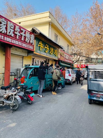 一张街头实拍照片，展示了一家名为「牛子炒鸡」的餐馆外景。黄色的两层建筑外挂满了红色和绿色的招牌，上面写着「家院牛子炒鸡」、「牛子炒鸡」等字样，并列出了干煸鸡、香辣鲤鱼、麻辣猪蹄等特色菜品及订餐电话。餐馆门口聚集着几位顾客，路边停放着蓝色三轮车、白色小轿车等车辆。背景中可以看到秋冬季节的树木，树叶呈现出黄色和棕色，天空晴朗湛蓝。