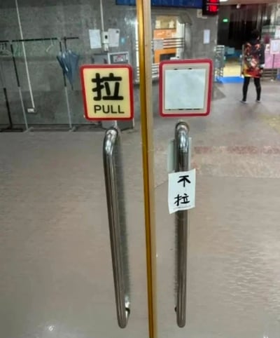 图片展示了一个公共室内场所（可能是地铁站或商场）的双开门场景。左侧门把手上方有一个官方标识牌，红底黄框，内部用黑色中文写着“拉”，下方配有英文“PULL”。右侧门把手上原本应有标识的区域为空白，但在门把手中下部贴着一张手写的白色纸条，上面用黑色马克笔写着“不拉”。背景中可见灰色墙壁、挂着的雨伞、一些设备以及远处一位行走的路人。
图片的幽默之处在于两侧门的指示信息相互矛盾，左侧指示“拉”，右侧却手写提示“不拉”，形成了“到底要不要拉门”的搞笑困惑场景。