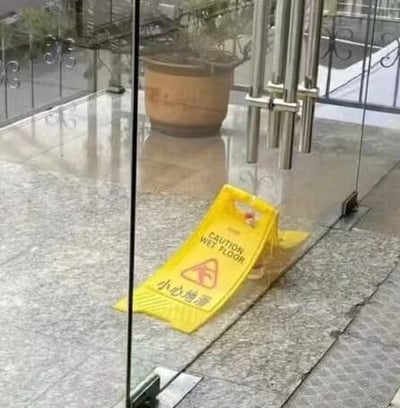 图片展示了一处玻璃门入口场景，地面铺有光亮的大理石地砖。门口地面上放置着一块黄色的“小心地滑”警示牌，牌上同时印有英文“CAUTION WET FLOOR”和中文“小心地滑”字样。背景中可见一个大型盆栽和透过玻璃门看到的室外区域，整体场景像是商场、办公楼等公共场所的入口处，旨在提醒行人注意地面湿滑。
