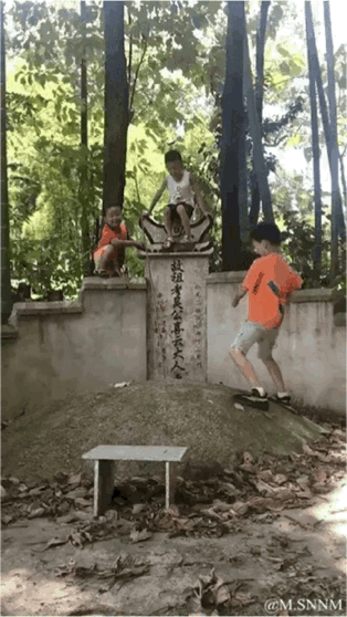 这是一段动态GIF，画面呈现了三个小男孩在一处林间墓碑区域玩耍的场景：一个男孩坐在墓碑的碑顶，姿态放松；一个男孩蹲在墓碑侧面的矮墙上；还有一个男孩正踩着墓碑的坟包边缘，似乎在移动。周围是茂密的树林，地面散落着落叶，这种行为既不尊重逝者，也存在跌落受伤的安全隐患，同时也反映出孩童缺乏相关礼仪和安全意识。