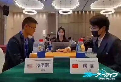 这是一场正式国际围棋赛事的对弈现场画面，画面右侧是中国围棋九段选手柯洁，他佩戴口罩，坐在桌前，左手放在桌上，姿态严肃专注；左侧是来自美国的漆圣颐初段，正目光看向柯洁，神态认真。两人面前的桌牌标注了各自的姓名、段位以及所属国家的国旗，桌上摆放着矿泉水，后方可见赛事工作人员和其他参会人员，右下角带有手游《蔚蓝档案（Blue Archive）》的水印，整体场景氛围正式严肃，是专业围棋赛事的现场。