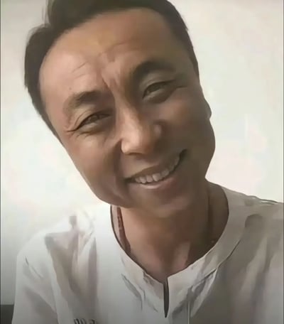 这是中文互联网知名梗人物神鹰哥（神鹰黑手哥）的经典近景大头素材照，图中神鹰哥身穿白色立领上衣，面向镜头咧嘴露笑，因为拍摄时的动态晃动，脸部呈现出明显的拉伸歪脸变形效果，这个特殊的视觉效果也成为该梗最具标志性的特征之一。