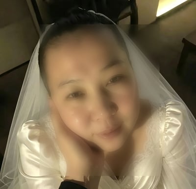 这是一张真人拍摄的静态自拍图，画面主体是一名东亚女性，她身着带有蕾丝装饰的白色泡泡袖婚纱，头上佩戴白色长款头纱，头发向后盘起，右手托住脸颊面向镜头，神情柔和带浅淡笑意，背景为光线偏昏暗的室内环境，画面整体带有柔焦效果。