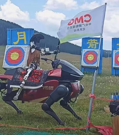 这张照片拍摄于户外射箭活动场地，背景是开阔的草甸、远处的山林与多云的天空。画面主体是一个装配机械义肢、头戴宽檐帽的人形骑手，骑在红黑配色的“骥翼T1000”四足仿生机械马上，机械马背还铺有带有民族风格红花纹样的鞍垫，骑手正手持弓箭做出瞄准动作。场地内排列着多个射箭靶位，靶位上方的蓝色箱体分别印有黄色的“月”“箭”字样，一侧旗杆上插着一面倒置的中央广播电视总台北京总站的旗帜，场地边缘还放置有收纳箭支的箭筒，整体呈现出未来科技感与传统射箭运动碰撞的新奇效果。