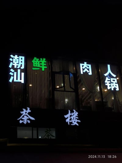 这是一张夜晚拍摄的线下餐饮店铺照片，店铺的窗户上安装了发光字招牌，从左至右为蓝白色的「潮汕」、绿色的「鲜」、蓝白色的「肉」「人锅」，组合起来应为「潮汕鲜肉火锅」（疑似字牌安装或显示失误），下方还有蓝白色的「茶楼」字样。透过打开的窗户和玻璃，能看到店内暖黄的灯光、窗帘以及模糊的店内用餐场景，整体氛围温馨，显示出该店铺同时经营潮汕风味火锅与茶楼两类业务。