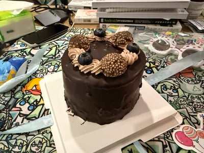 一张巧克力生日蛋糕的特写照片，蛋糕表面覆盖着光滑的巧克力涂层，顶部装饰有棕色巧克力球、蓝莓和奶油花。蛋糕放置在白色方形蛋糕纸上，下方是印有彩色动漫人物图案的桌布。背景中可见一些日常物品，包括手机、书籍、纸巾盒等，营造出温馨的生活氛围。