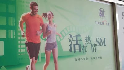 天津SM广场的户外广告牌，画面主体为两名正在慢跑的年轻人（男性穿橙色T恤黑色短裤，女性穿灰色背心灰色短裤），两人面带笑容，展现活力满满的运动状态。背景为绿色渐变，融入城市建筑剪影元素。右侧有“SM”品牌标识、“TIANJIN 天津”字样及中文标语“活热SM 活在情”“运动小装备，健康大事情”，整体传递积极健康的生活理念。