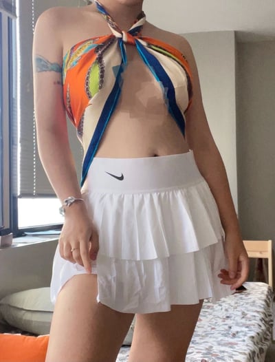 一位女性站在室内房间中，上身穿着由彩色丝巾系成的挂脖上衣，丝巾上有橙色、蓝色、绿色等多种颜色的图案，领口处打有蝴蝶结。下半身穿着一条白色的Nike运动短裙，裙子有多层百褶设计。她的左臂上有一处纹身，手腕上戴着手表和戒指。背景中可以看到窗户、床铺和椅子等家具，整体呈现出日常居家的穿搭场景。