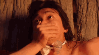 动态图展示了一位身处原始部落木屋内的 shirtless 男子，他佩戴着项链，表情夸张地用手捂住嘴巴，似在大笑或极度震惊。画面中还出现其他部落成员躺卧的场景，以及燃烧的火焰，整体营造出原始、粗犷的氛围，男子的夸张反应是视觉焦点，常被用作表达强烈情绪的反应素材。