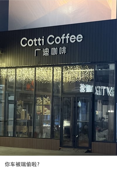 这张图为实拍的咖啡店门店外观，原本的“库迪咖啡”招牌的“库”字缺失了“车”的部分，仅剩“广”字，变成了“广迪咖啡”，上方的英文“Cotti Coffee”字样完好无损，门店玻璃内可以看到装饰的串灯和店内陈设，图片底部配有文字“你车被瑞偷啦？”。该图玩了文字拆解的趣味梗：“库”字去掉“车”即为“广”，配文调侃库迪的“车”是被竞品瑞幸偷走了，笑点结合了库迪与瑞幸的品牌竞争背景，十分幽默。