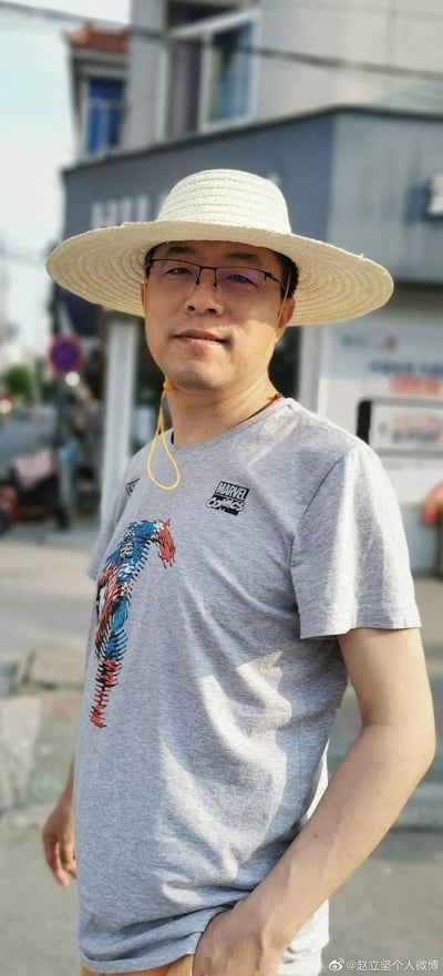 这是一张赵立坚的户外实拍照片，他头戴浅米色宽檐草帽，搭配黑框眼镜，面带温和的微笑看向镜头。身上穿着一件灰色短袖T恤，T恤左胸位置印有黑色的“MARVEL COMICS”标识，衣身还有由红蓝配色构成的美国队长元素图案。他一只手插在裤袋里，姿态放松自然，背景是普通的临街街道与建筑，整体风格十分接地气，和他以往的官方工作形象形成鲜明反差，展现出生活化的一面。