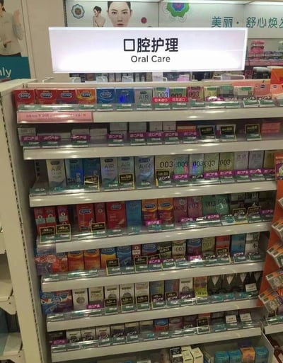 超市的口腔护理货架区域，上方悬挂着写有“口腔护理 Oral Care”的白色标牌，货架上却主要摆放着各类避孕套产品。可见多个品牌如杜蕾斯（Durex）、AIR以及标注“003”“日本进口”的避孕套包装盒，颜色多样，价格标签显示部分商品售价¥9.9至¥288不等，还标有“进口产品”“买一送一”等促销信息。传统口腔护理用品如牙膏、牙刷等在货架上占比极少，形成明显的商品分类错误，私密用品与日常口腔护理产品意外同区陈列，场景既滑稽又令人啼笑皆非。