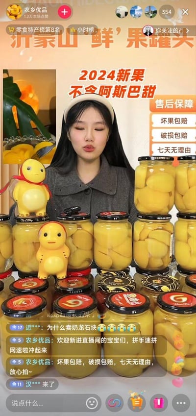 这是一张电商直播带货的截图，画面中央是一位女性主播，她面前摆放着多罐沂蒙山鲜果罐头，罐头顶部还放置了可爱的奶龙玩偶作为装饰。图片上方有醒目的促销标题“2024新果 不含阿斯巴甜”，突出产品卖点。右侧列出了售后保障政策，包括坏果包赔、破损包赔和七天无理由退货。左下角显示了观众评论和主播回复的互动内容，整体氛围热闹且具有吸引力，旨在促进产品销售。