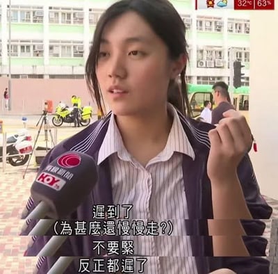 图片为一则新闻采访场景，一名穿着紫色校服外套和白色条纹衬衫的女生正在接受采访，面前的麦克风上有“HOY TV 香港開電視”标识。背景可见街道、建筑物、摩托车及行人。女生表情淡定，右手握拳置于身前。图中字幕显示对话内容：“遲到了（為甚麼還慢慢走？）不要緊反正都遲了”，展现出一种对迟到满不在乎的幽默态度。