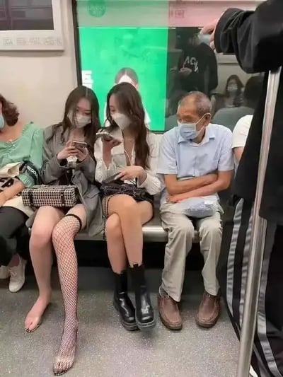 这张图片拍摄于地铁车厢内，画面中左侧有两位穿搭时尚的年轻女性和一位老年男性一起坐在座位上，三人的气质和穿着形成鲜明对比。左侧女生穿着镂空网袜和时尚外套，另一位女生则是短裙靴子，两人低头专注玩手机。老年男性则穿着普通的衬衫和长裤，双手抱着袋子坐姿拘谨，显得有些局促。整个画面展现了地铁这样公共空间里不同年龄层的生活状态、穿衣风格和气氛反差。这类画面在网络上容易引发关于“代际差异”、“穿衣自由”和“社交场合礼仪”的讨论。无明显双关、谐音梗，但对比感突出带来轻微幽默感。
