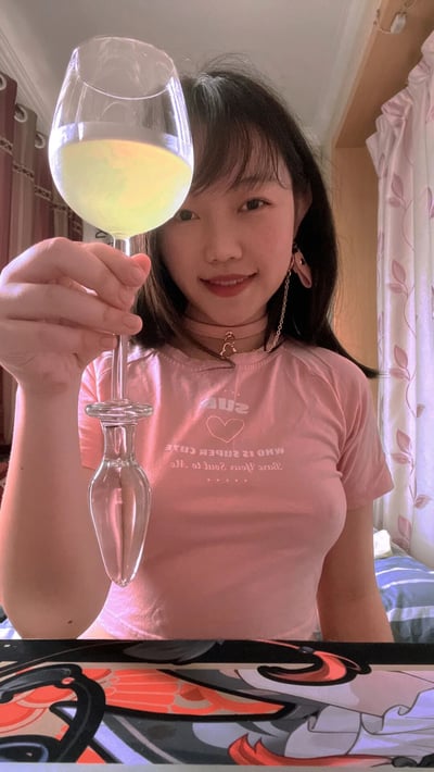一位年轻女性在室内环境中手持装有黄色液体的高脚杯自拍，面带微笑。她穿着粉色T恤，上面印有"SUGAR WHO IS SWEETER THAN ME"字样，佩戴耳环和项链，背景可见带有花纹的窗帘和床铺。