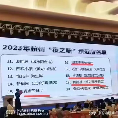 这是一张活动现场的实拍图，展示了2023年杭州“夜之味”示范店名单的第11至20位入选店铺，其中麦当劳、肯德基的多家门店在列，且部分麦当劳、肯德基门店被红色下划线标注。画面中可见台前的工作人员与台下的参会人员，场景为正式的名单发布或颁奖类活动，呈现了杭州夜经济相关的餐饮评选结果。