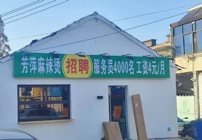 这是一张拍摄于街边的原始照片，主体是一家名为“芳萍麻辣烫”的小型店铺，店铺外墙为白色，屋顶是斜顶结构，门口摆放着未拆封的纸箱，看起来可能是刚开业或正在整理店内物品。店铺上方悬挂着一块绿底招牌，左侧标注店名“芳萍麻辣烫”，中间黄框区域写着“招聘”字样，右侧则是“服务员4000名 工资4元/月”的招聘内容。这个信息存在明显的乌龙：一家街边小型麻辣烫店不可能需要4000名服务员，且月薪4元远低于正常薪资标准，大概率是排版失误，比如将“4名服务员，工资4000元/月”的内容错误拆分排版，或是输入时遗漏数字，从而形成了荒诞搞笑的效果。
