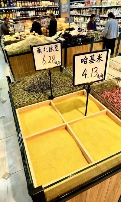 超市粮食区的场景，多个透明玻璃柜中摆放着不同种类的粮食。最显眼的是两个价格牌，一个写着'南北绿豆 6.28元/斤'，另一个则幽默地写着'哈基米 4.13元/斤'。背景中可以看到几位顾客正在挑选商品。这是一个利用网络流行语'哈基米'（源自《间谍过家家》中安雅对狗的称呼）制作的搞笑梗图，将虚构的'哈基米'当作一种粮食来标价。