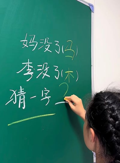 整张图是实拍静态内容，画面右下角可见一位梳着麻花辫的女性的侧后方，她正手持黄色粉笔在绿色的家用小黑板上书写，黑板上用白色粉笔写着拆字字谜：第一行是“妈没了(马)”，第二行是“李没了(木)”，第三行是“猜一字？”，其中括号里的“马”“木”、问号以及答案填写横线是用绿色粉笔书写。该字谜的解法为：“妈”去掉“马”字旁得到“女”字，“李”去掉“木”字头得到“子”字，二者组合起来就是“好”字，呼应了发布者的标题。另外谜面“妈没了马”的发音和“骂”读音相近，因此衍生出“被骂了两遍还要说好”的趣味笑点。