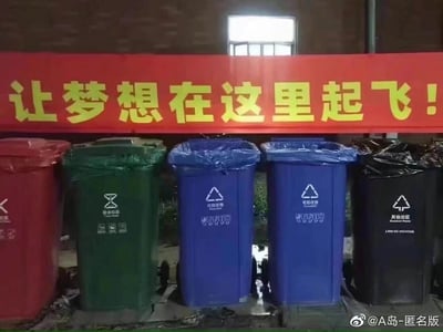这是一张充满反差感的现实场景图，画面上方悬挂着一条红色底黄色字体的励志标语“让梦想在这里起飞！”，标语下方整齐摆放着五个分类垃圾桶：从左至右依次是套着红色垃圾袋的有害垃圾桶、套着绿色垃圾袋的厨余垃圾桶、两个套着蓝色垃圾袋的可回收物垃圾桶、套着黑色垃圾袋的其他垃圾桶，垃圾桶外观规范，带有对应的分类标识。励志标语的宏大感与下方垃圾桶的日常、“归宿感”形成强烈反差，带有戏谑意味，仿佛在调侃理想与现实的落差，传递出一种对生活或工作中梦想落空的无奈情绪。