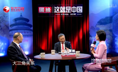 这是东方卫视《这就是中国》节目的一个访谈场景，画面中有三个人围坐在圆形桌子旁进行讨论。左边是一位戴眼镜的男士，穿着深色西装；中间是一位系着红色领带的男士，正手持物品发言；右边是一位穿着粉色套装的女士，手持麦克风。桌子上摆放着带有节目标识的杯子和物品。背景是红色幕布，两侧屏幕分别展示了天坛和长城的图像。屏幕底部出现“大三角关系”的字幕，显示他们正在讨论相关话题。
