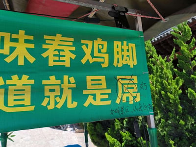 这是一张户外拍摄的小吃摊招牌照片，招牌为绿色底搭配黄色醒目大字，上方写着经营品类“味舂鸡脚”，下方原本的“道就是屌”中，“屌”字因用词不文明被人用黑笔圈画打叉，旁边手写替换为“好”字，同时在招牌下方添加了“文明景区，从我做起”的手写文字，是景区文明建设中对不文明标语进行修改的真实街头场景，充满接地气的生活感。
