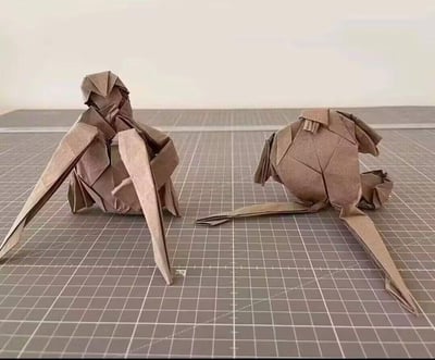 这张图展示了两个用牛皮色纸张制作的创意折纸作品，放置在带有白色网格的灰色切割垫上。左侧是猴子的上半身造型，有着明确的头部、手臂结构，两只手臂向前伸展呈支撑姿态；右侧是猴子的下半身，带有尾巴和腿部结构，二者被拆分开摆放，仿佛一个完整的猴子被分成了两部分，尽显折纸工艺的精细与创意构思的巧妙。