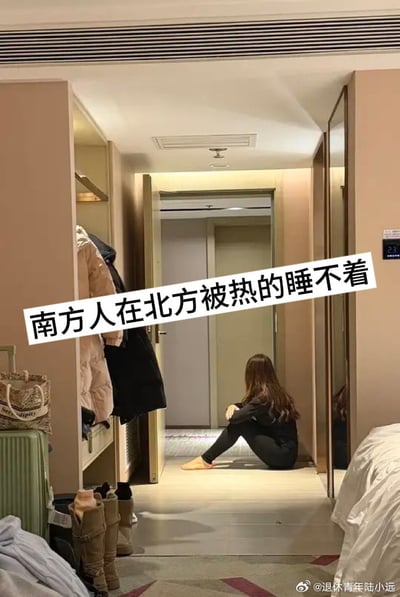 图片展示了一个室内走廊场景：左侧是挂有外套的开放式衣柜，右侧可见部分床铺，中央走廊地面上坐着一位长发女子，她身着黑色衣物、赤脚，背对着镜头望向远处的房门。画面中央有醒目的白色文字覆盖：“南方人在北方被热的睡不着”，生动呈现了南方人到北方后因不适应暖气过足而难以入睡的有趣生活体验，通过真实场景与文字结合，幽默展现了南北气候适应差异的普遍现象。