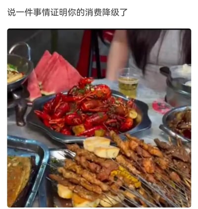 图片上方配有文字“说一件事情证明你的消费降级了”，下方是一张摆满丰盛食物的餐桌照片，包括一大盘红彤彤的小龙虾、多串烤肉烤串、切块西瓜以及其他菜肴，营造出热闹的聚餐氛围。图片通过“文字提问”与“画面呈现”的反差制造幽默效果，暗示“在家吃大餐”成为替代外出消费的“降级”方式，是对当下消费观念变化的调侃。