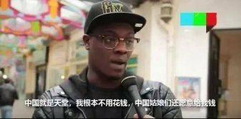 这是一张街头采访的视频截图，图中的黑人男子戴着黑色字母帽子与眼镜，正对着麦克风接受采访，背景是挂满彩色装饰的街道。画面下方配有中文字幕：“中国就是天堂，我根本不用花钱，中国姑娘们还愿意给我钱”，属于真实采访场景被添加中文字幕后的传播内容，表述具有一定争议性。