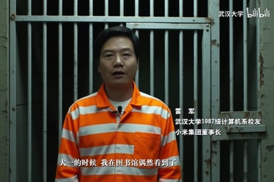 这是哔哩哔哩平台上武汉大学相关视频的截图，小米集团董事长、武汉大学1987级计算机系校友雷军身着橙白条纹的类似囚服的服装，站在铁栅栏门前，正对着镜头讲述自己大一在图书馆的经历。该造型为节目创意设计，并非真实入狱场景，用于分享个人大学往事。