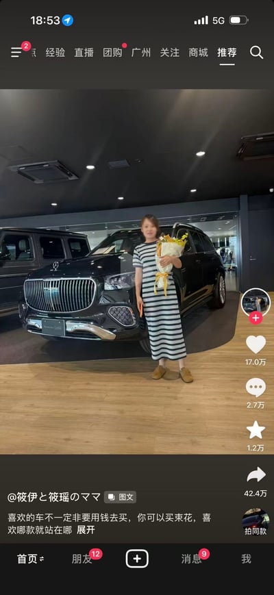 这是一张抖音平台的内容截图，拍摄场景为汽车4S店展厅，一名身穿黑白条纹长款连衣裙、脚穿棕色平底拖鞋的女士面带微笑，手持包装好的黄色花束站在一辆黑色迈巴赫SUV车头旁，车辆后方还停放着一辆黑色奔驰G级越野车。该条内容的发布者配文称“喜欢的车不一定非要用钱去买，你可以买束花，喜欢哪款就站在哪”，用调侃的方式表达无需实际购车，只要站在喜欢的车旁拍照就相当于“拥有”了车辆。右侧显示该内容获得17.0万点赞、2.7万条评论、1.2万次收藏、42.4万次转发，界面顶部显示当前定位为广州，底部是抖音的常规导航栏。