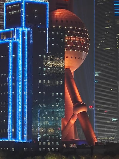 这张图片展示了上海标志性建筑东方明珠塔的壮丽夜景。东方明珠塔位于画面中央，其独特的球形结构被温暖的橙色灯光照亮，球体表面布满了小星星般的光点。塔的底部由粗壮的支柱支撑，呈现出稳定而宏伟的姿态。周围环绕着现代化的摩天大楼，左侧建筑被蓝色灯光勾勒出几何轮廓，右侧则是高耸的玻璃幕墙大厦。整个画面展现了上海繁华的都市夜景，灯光璀璨，彰显出现代大都市的活力与魅力。