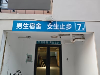 图片展示了一栋宿舍楼的入口处，上方悬挂着蓝色的标识牌，写有'男生宿舍 女生止步 7栋'，而在其正下方的门上又有一块绿色标识牌，写着'女生宿舍 男生止步'。两块标识牌内容相互矛盾，形成了明显的信息冲突，可能是管理疏忽导致的乌龙场景。