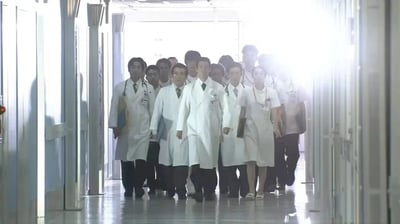 一群穿着白大褂的医护人员正列队行走在医院的走廊中，队伍前方有几位医生领头，后方跟随其他医护人员。走廊两侧是白色的墙壁和病房门，地面光洁反光。从画面后方照射过来的强烈光线在人物身后形成了明显的光晕效果，营造出一种专业且略带神圣感的氛围。整个场景给人一种医疗团队正在紧急赶往救治现场或进行重要工作的印象。