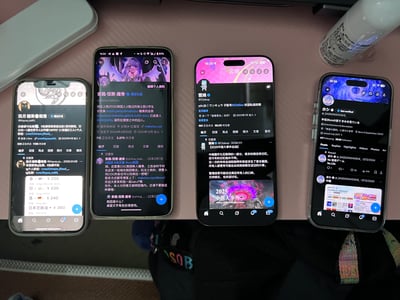 桌面上并排放置四部智能手机，屏幕均亮着显示不同社交平台的个人主页。手机从左到右依次为：黑色猫头像账号界面、紫色系虚拟角色“紫晶·恒野·霞”的粉丝页（3.87万粉丝）、粉色主题“雲鸠”账号主页（含2025中推大作宣传图）、蓝色系日文账号界面。背景可见桌面物品如白色容器和瓶装物品，整体呈现日常桌面多设备展示社交账号的场景。