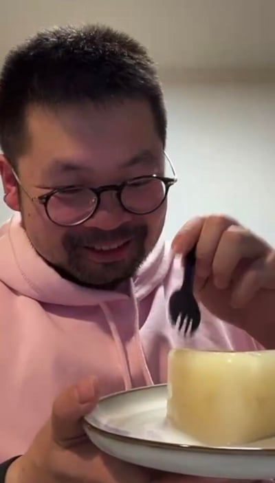 一位戴着黑框眼镜、留着胡须的男士，身穿粉色连帽卫衣，正笑着用黑色小叉子准备享用盘子里的浅黄色甜点，看起来心情十分愉悦，画面给人一种轻松温馨的日常感。