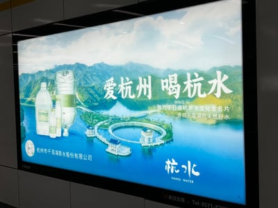 这是一张地铁站内的杭州市地方品牌广告，主推饮用水品牌“杭水”。画面左侧为几种不同瓶型的瓶装水，正中央和右侧为千岛湖和周边自然风景，突出水质纯净和自然来源。广告语“爱杭州 喝杭水”呼应了城市文化和水源地的特色。整体氛围清新自然，视觉主体突出，强调地方品牌和自然环保理念。

本广告未涉及任何双关、谐音梗或暗示内容，定位清晰，强烈地方认同感和健康生活理念。