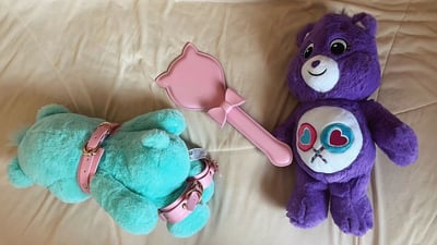 画面中展示了两只Care Bears爱心熊玩偶和一个粉色猫咪造型paddle的创意摆放场景。左侧是一只蓝绿色爱心熊，呈趴卧姿势，颈部和腿部各佩戴了一条粉色皮带；右侧是一只紫色爱心熊，直立坐着，腹部有两个交叉的彩色爱心图案，面带微笑；两者之间放置着一个粉色猫咪形状、带蝴蝶结手柄的paddle。背景为米色床单，整体氛围可爱中带着一丝反差萌的幽默。
