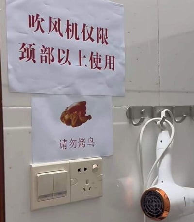 这是在贴有白色瓷砖的墙面上的实拍场景，上方贴有红色字体的提示标语“吹风机仅限颈部以上使用”，下方搭配着一张烤鸡的图片，配文“请勿烤鸟”，旁边挂钩上挂着白色吹风机，下方是开关插座。该标语是为了规范吹风机的使用，调侃有人用吹风机烤鸟的离谱行为，用烤鸡图来暗示烤鸟后的结果，风格搞笑接地气。