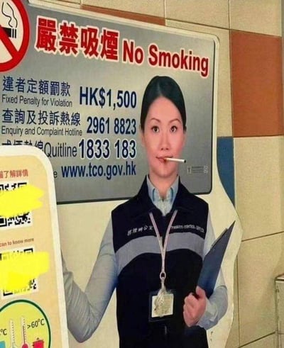 这是一张经过P图改编的静态单图，画面呈现的是香港公共场所的一处禁烟宣传场景：背景是带有中英文的“严禁吸烟”标识牌，上面标注了违例罚款1500港币、查询投诉热线、戒烟热线以及相关官方网址；前景是一个港铁工作人员的立牌，原本应该配合禁烟宣传的立牌被网友恶搞，在其嘴部P上了一支点燃的香烟，形成了强烈的反差感，让原本严肃的禁烟宣传场景变得滑稽搞笑，充满了恶搞趣味。